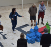 Groupe de touristes en visite guidée dans la cours du Belvédère. Une guide montrant, au sol, les formes des bâtiments de la ville de Saint-Benoît-sur-Loire et la maquette miniature de l'Abbaye de Fleury